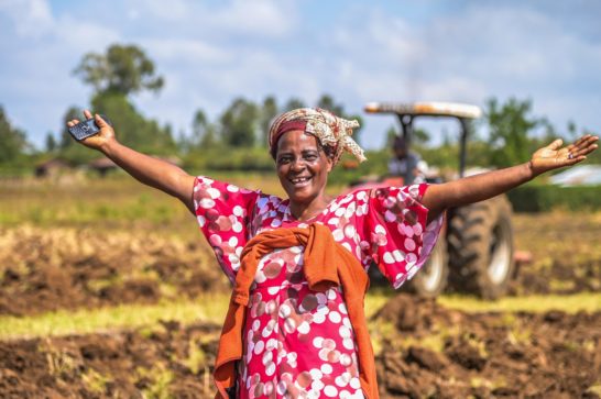 smiling woman standing in a farm plot holding a mobile and spreading both arms into the air