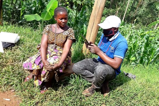 woman on the left and man next to her sitting in the grass the man holding a tablet and taking notes lush corn field in the background