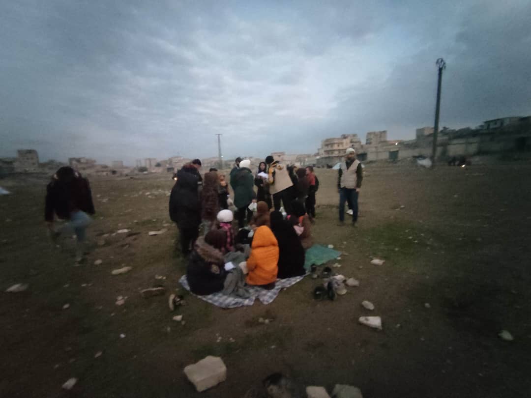 stony field next to houses in ruins with people carrying bags