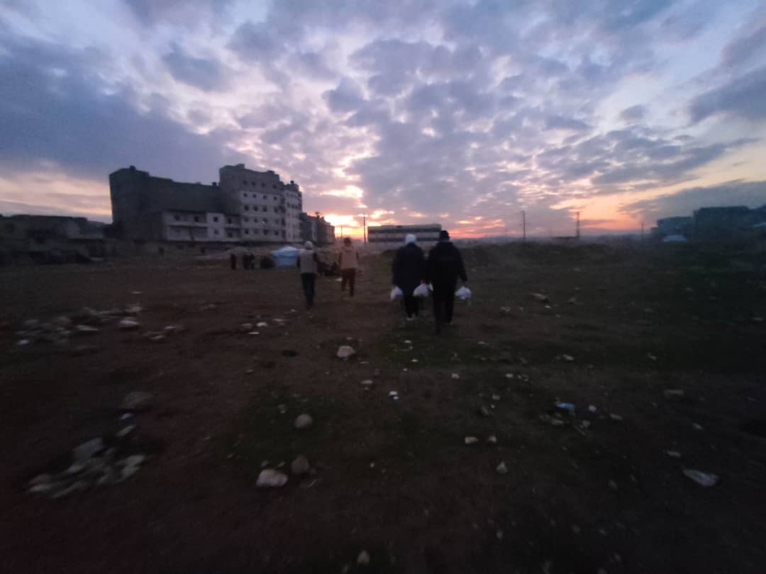 stony field next to houses in ruins with people carrying bags