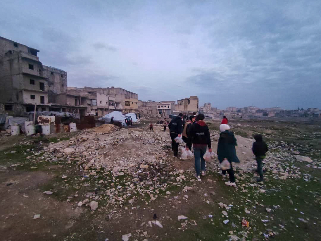 stony field next to houses in ruins with people carrying bags