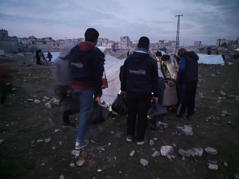 stony field next to houses in ruins with people carrying bags