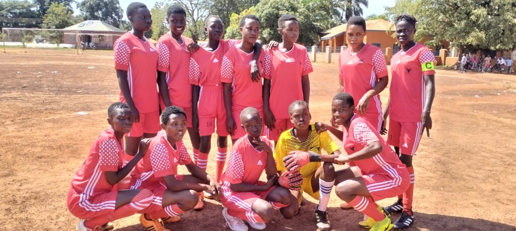 a girls football team standing on a sandy pitch wearing football outfits