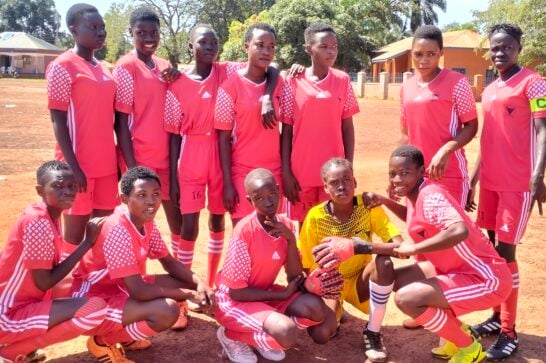 a girls football team standing on a sandy pitch wearing football outfits