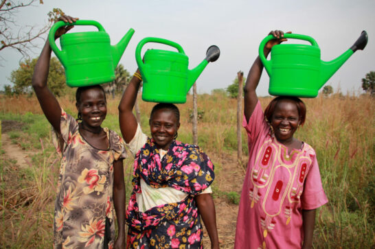Three women smiling holding bright green watercans on their heads