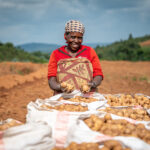 Een lachende Burundese boerin staat op een aardappelveld met gevulde zakken voor zich. Ze houdt een paar aardappelen in haar handen en draagt een rode blouse en hoofddoek. Op de achtergrond zijn heuvels en akkers te zien onder een licht bewolkte lucht.