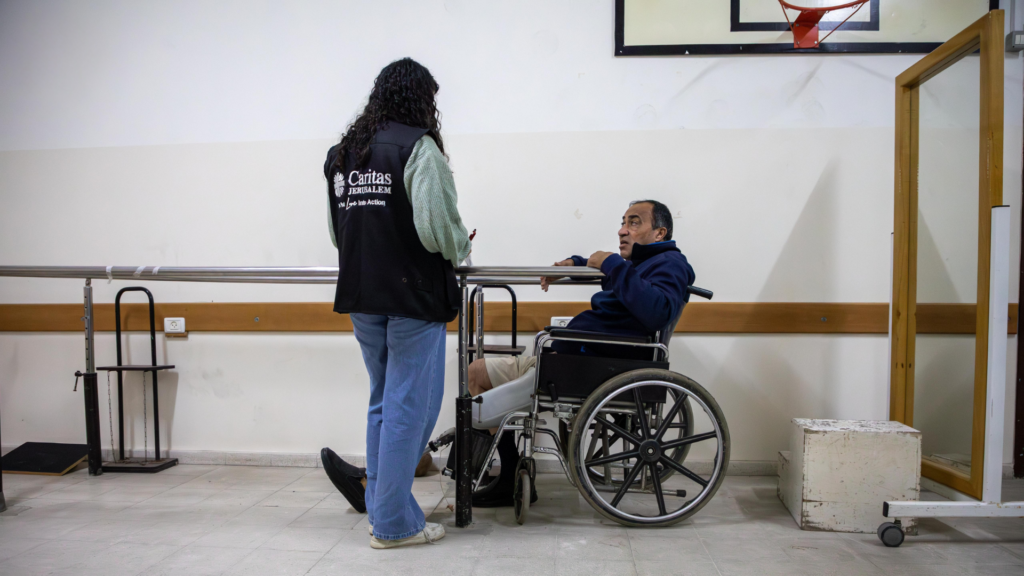 Een man in een rolstoel voert een gesprek met een hulpverlener van Caritas Jerusalem, die met haar rug naar de camera staat. Ze bevinden zich in een revalidatieruimte met een looprek en wandbeugels.