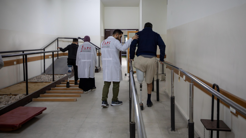 Revalidatiepatiënten met een beenprothese oefenen het lopen in een therapieruimte met verschillende ondergronden en trapjes. Ze worden begeleid door fysiotherapeuten in witte jassen van het ALPC-centrum in Gaza.