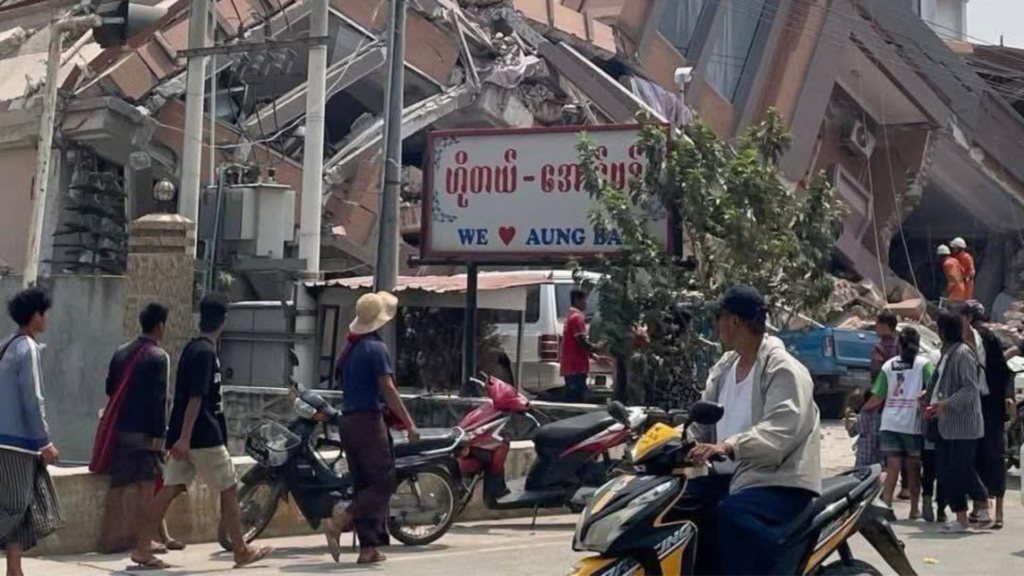 Mensen staan en rijden op scooters voor een ingestort gebouw in Myanmar, na een aardbeving; op de achtergrond zijn reddingswerkers zichtbaar tussen het puin.