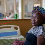 Zwangere vrouw met gekleurde doek op hoofd zit in Harare Zimbabwe op een bed in een kliniek en kijkt in camera.