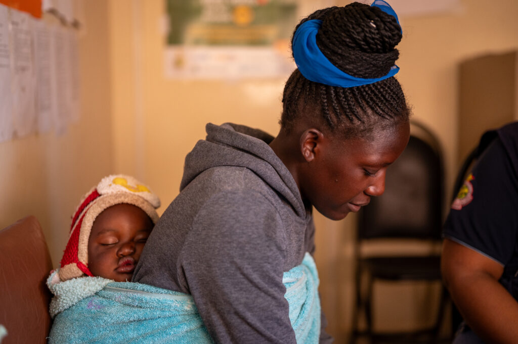 Zimbabwaanse vrouw zit met baby op haar rug in wachtkamer van een kliniek in Harare.
