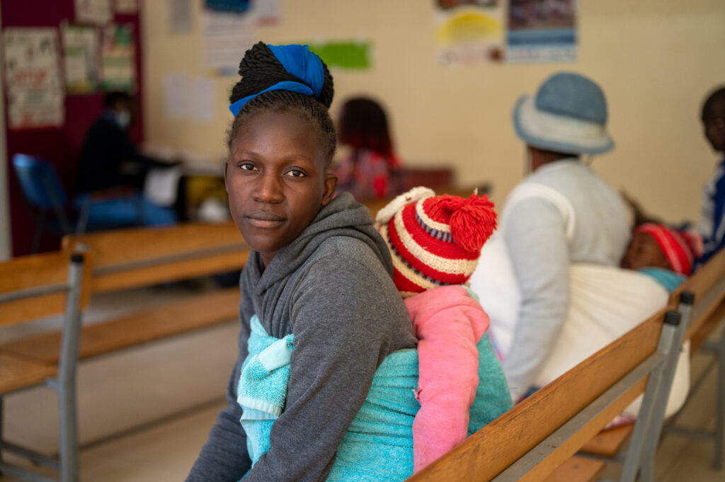Zimbabwaanse vrouw zit op een bank in een kliniek met haar baby op haar rug. Op de achtergrond zit een andere moeder met baby op haar rug.