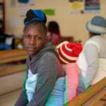 Zimbabwaanse vrouw zit op een bank in een kliniek met haar baby op haar rug. Op de achtergrond zit een andere moeder met baby op haar rug.