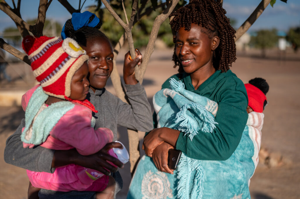 Twee Zimbabwaanse vrouwen die zorgvoucher van Cordaid hebben ontvangen staan buiten en lachen naar de camera. Een heeft haar baby op haar rug, de ander in haar armen.