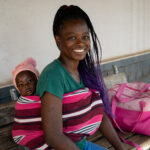 Trish, een van de deelnemers van het voucherprogramma van Cordaid, met dochter Adèle op rug op een bankje voor de Mbare kliniek in de sloppen van Harare, Zimbabwe.