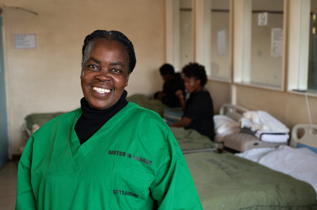 Zimbabwaanse verloskundige en hoofdverpleegkundige Juliette lacht in de camera. Op de achtergrond liggen twee zwangere vrouwen in hun ziekenhuisbed.