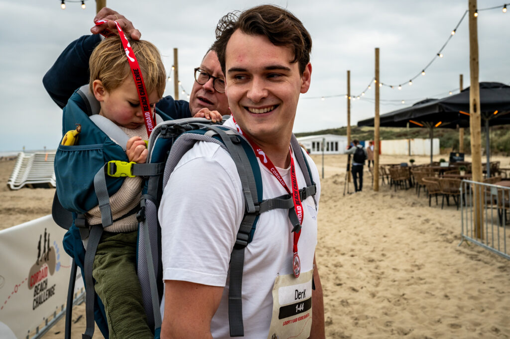 Vader met zoon op rug bij finish Cordaid Beach Challenge 2025, krijgen medaille van presentator Edwin Koene